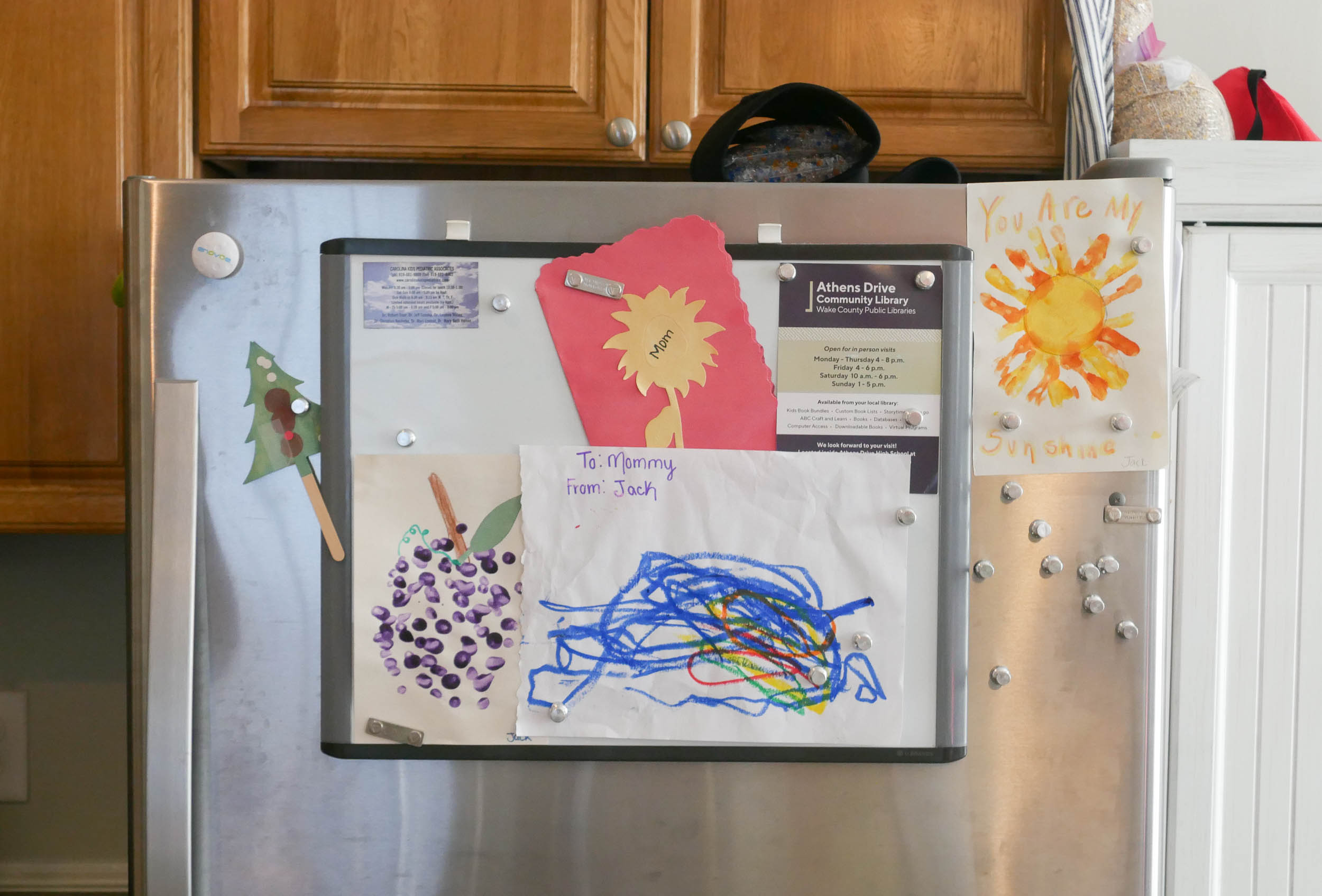 The top half of a refrigerator is covered with several pieces of a child’s art. A painted image of a yellow sun reads, “You are my sunshine.” An abstract, colorful scrawl reads, “To Mommy From Jack.” 