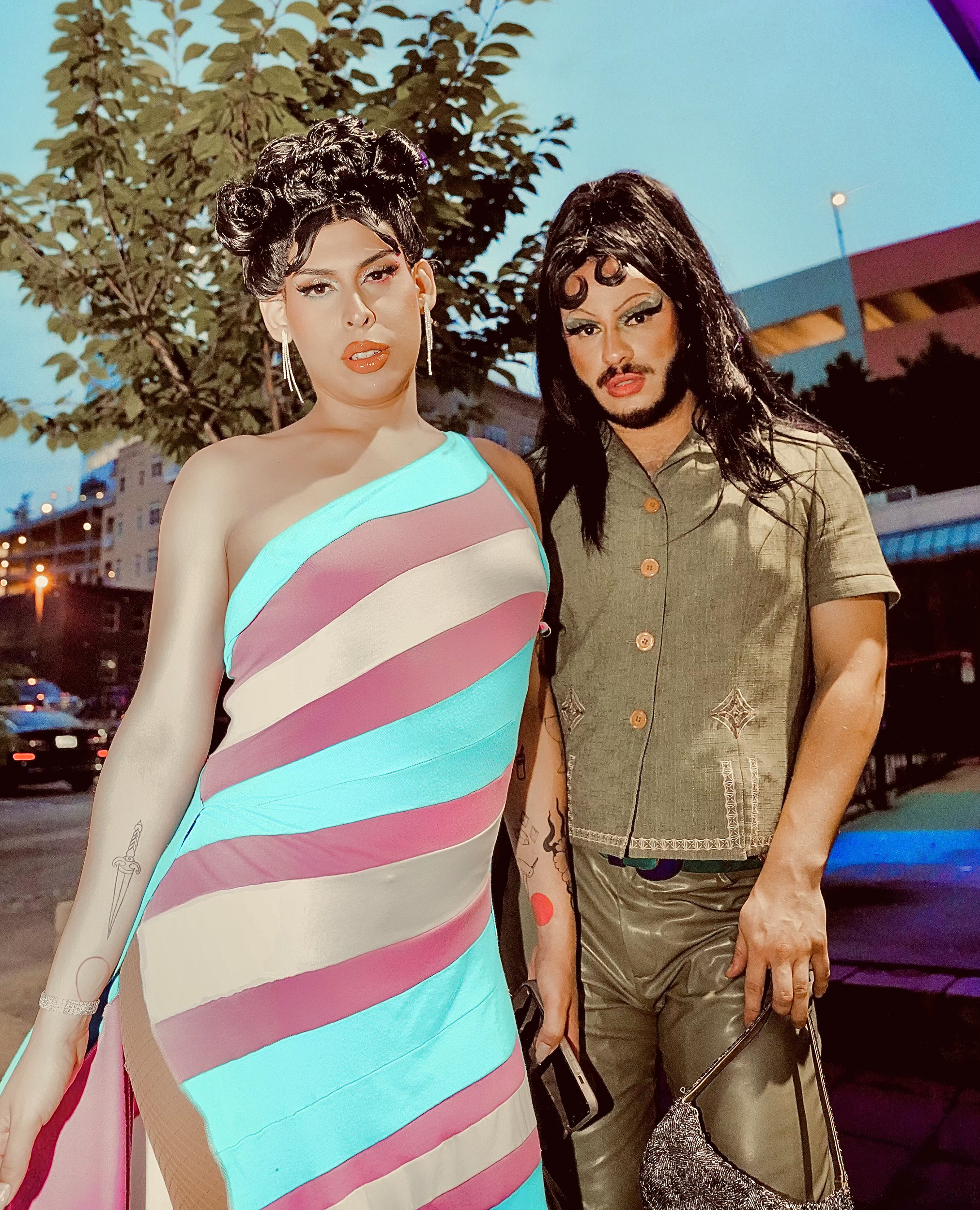Kali Fuchis and Miss B. Haven pose together in full drag, Fuchis is wearing a green textured shirt with leather pants and Miss B. Haven is wearing a blue and pink-striped dress.